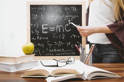 Woman writing physics equations on a blackboard with books and an apple on the desk.